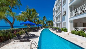 A poolside area with lounge chairs and umbrellas, a clear view of the ocean, and a building with balconies.