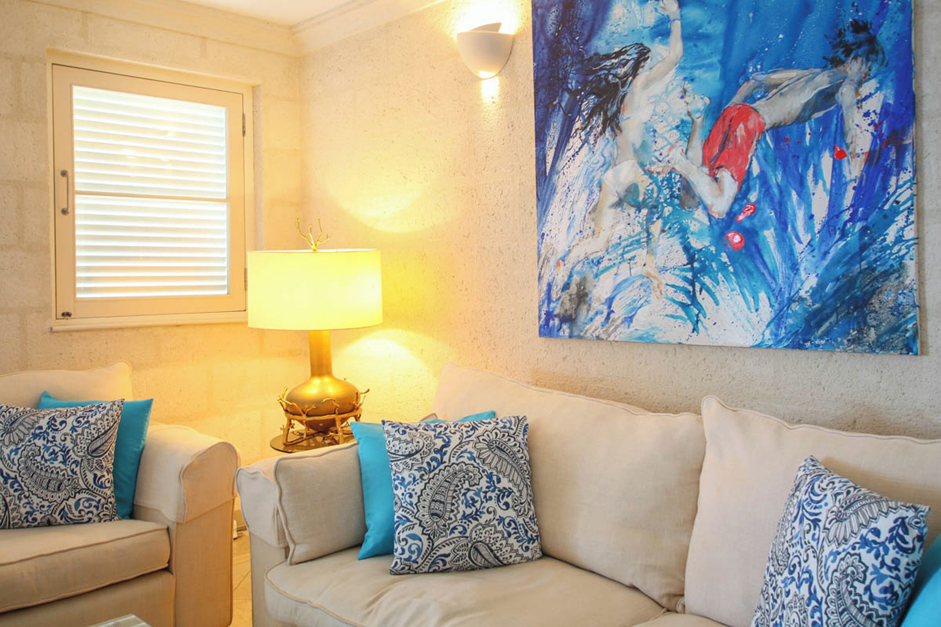 A living room with a beige sofa, patterned cushions, a table lamp, and a painting on the wall.