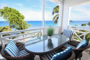 Ein Balkon mit Korbmöbeln und einem Glastisch, mit Blick auf einen Strand mit Palmen und den Ozean.