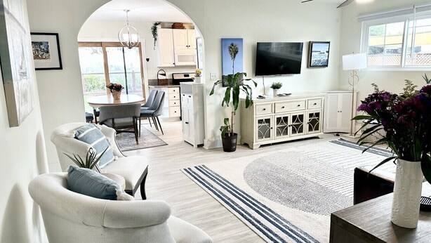 A modern living room with a dining area, white furniture, a flat-screen TV, and a ceiling fan. - Art Gallery Family Home - Southern California Style - Excellent Location (San Diego)