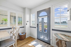 Una habitación con piso de madera, una mesa de comedor con sillas, una cesta de mimbre, una puerta con vistas y una mesita con una tabla de surf.