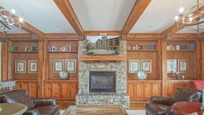 A cozy living room with a stone fireplace, leather sofas, wooden bookshelves, and a view of a building through a large window. - Highland House #102-104: 2Br: 2 Bath (Snowshoe)