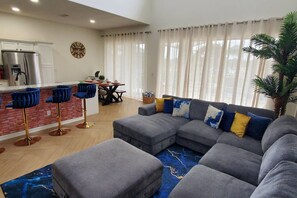 Una sala moderna con un sofá seccional gris, una zona de comedor con una mesa y sillas, una cocina con refrigerador, y un ventanal con cortinas.