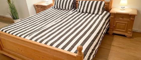 A wooden bed with a striped bedspread and matching pillows, flanked by nightstands with lamps, in a room with wooden flooring and a potted plant.