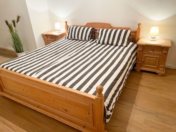 A wooden bed with a striped bedspread and matching pillows, flanked by nightstands with lamps, in a room with wooden flooring and a potted plant.