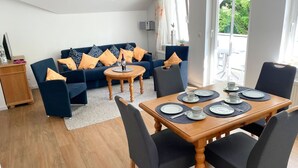 A modern living room with a sofa, chairs, a dining table set for four, and a view of the outdoors. - Villa Amanda - Strandnahe Fewo in Zentraler Aber Ruhiger Lage in Binz - Villa Amanda Fewo Nr.09 (Binz)