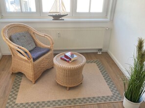 A wicker chair with a cushion and a matching ottoman, both placed on a patterned rug. A small sailboat model is visible on a windowsill above.