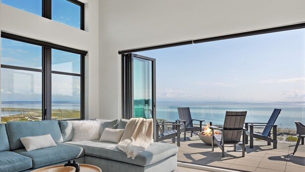 A modern living room with a large window overlooking a beach. There is a sofa, a coffee table, and a fire pit with chairs outside.