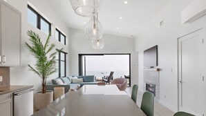 A modern kitchen with a large island, a dining area with green chairs, a fireplace, and a large window overlooking the ocean.