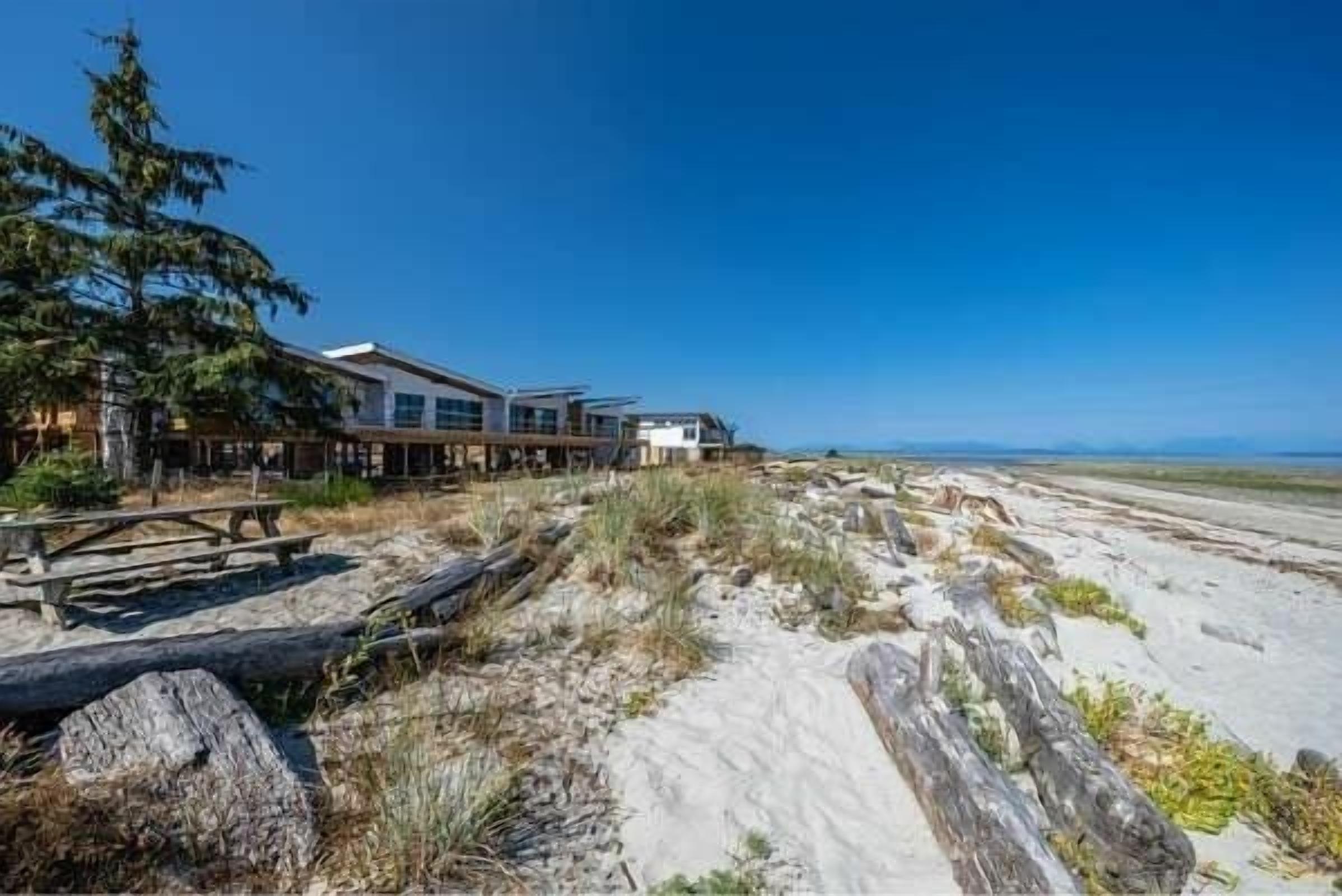 Ein Küstenhaus mit einer Holzterrasse, großen Fenstern und einem sandigen Strand im Hintergrund.