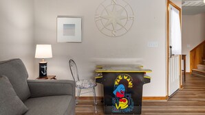 A living room with a grey sofa, a lamp, a chair, a wall-mounted compass, and a small table with a Pac-Man design. - Sunshine Daydream - Oceanside, Private Pool, Hot Tub and Game Room (Corolla)