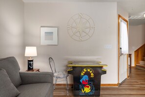 A living room with a grey sofa, a lamp, a chair, a wall-mounted compass, and a small table with a Pac-Man design. - Sunshine Daydream - Oceanside, Private Pool, Hot Tub and Game Room (Corolla)