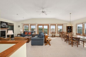 A spacious living area with a fireplace, comfortable seating, a dining table, and large windows offering views. - Sunshine Daydream - Oceanside, Private Pool, Hot Tub and Game Room (Corolla)