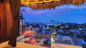 A room with a thatched roof, a view of a coastal town, and string lights. - Tree-House Apt with Panoramic Ocean View+Pool+Steps to Beach & Malecon/Boardwalk (Puerto Vallarta)