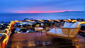 A wooden deck with a wicker chair and a small table, overlooking a coastal cityscape during sunset. - Tree-House Apt with Panoramic Ocean View+Pool+Steps to Beach & Malecon/Boardwalk (Puerto Vallarta)