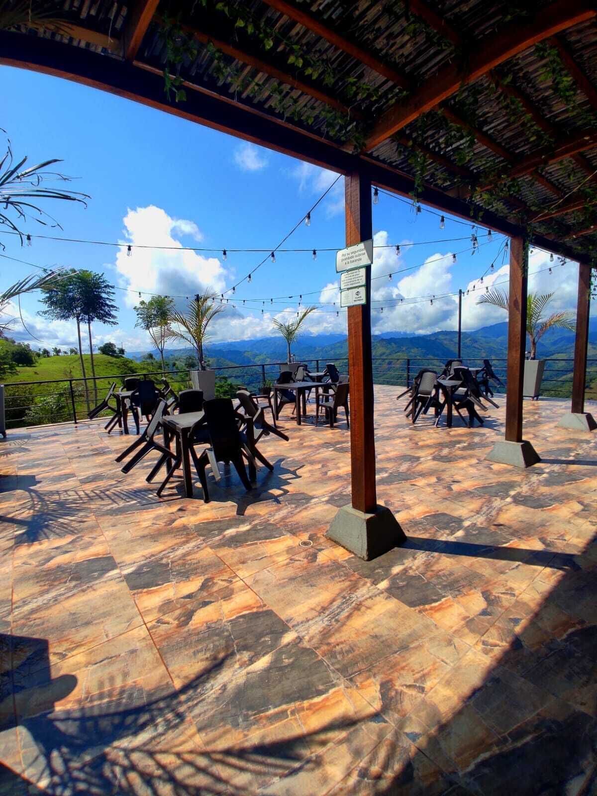 An outdoor seating area with tables and chairs, a pergola with string lights, and a view of mountains and greenery.