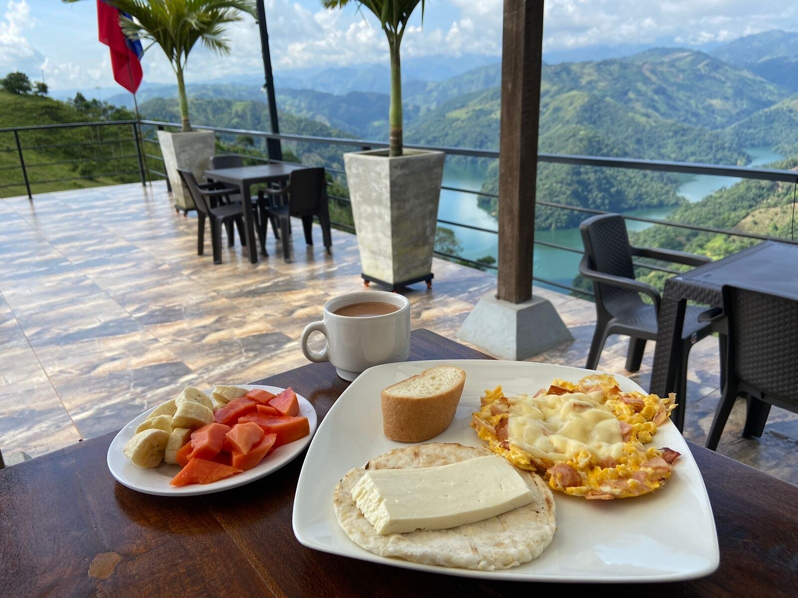 A breakfast setting with a scenic view, including a plate with toast, eggs, and fruit, and a cup of coffee.