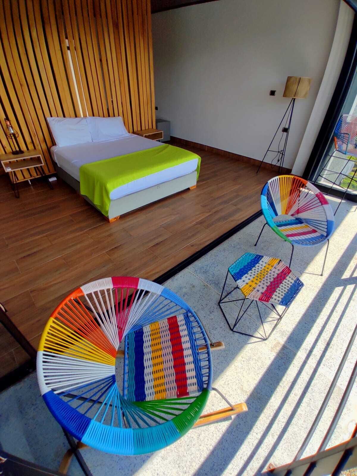 Un dormitorio moderno con una cama, un cabecero de madera y una manta verde. Hay dos sillas coloridas y una mesita.