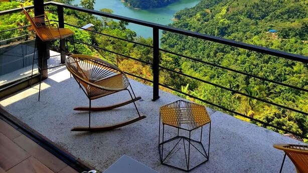Varanda com cadeiras de vime e mesa, com vista para uma paisagem montanhosa e um lago.