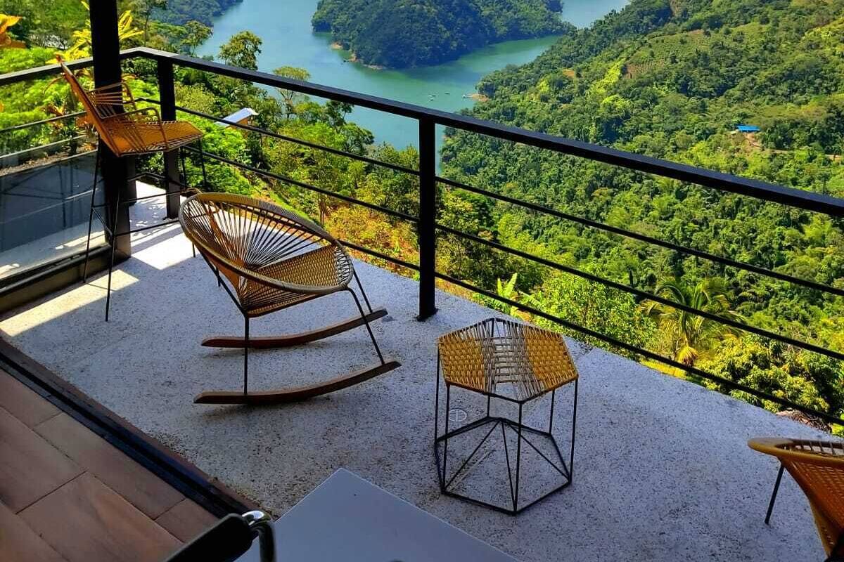 Een balkon met rieten stoelen en een tafel, met uitzicht op een bergachtig landschap met een meer.