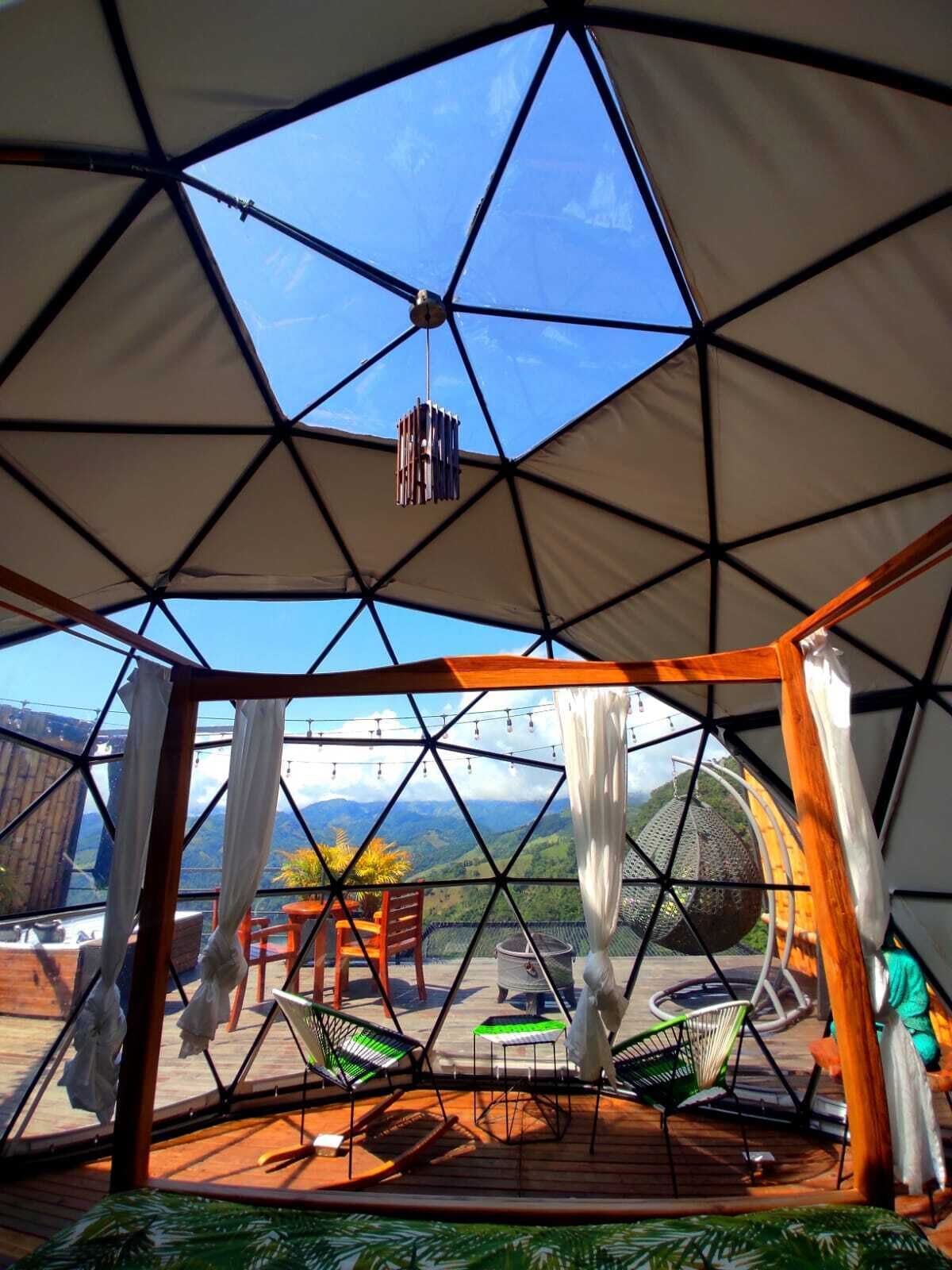 A geodesic dome structure with a view of a mountainous landscape. Inside, there are wooden tables and chairs, and a hanging light fixture.