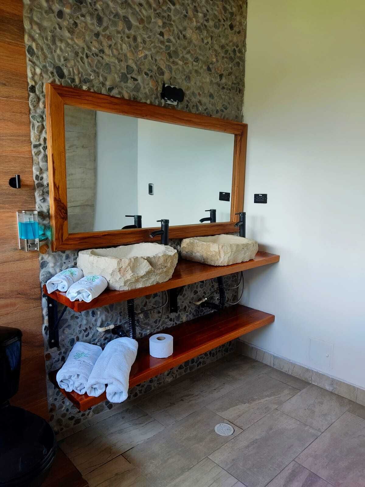 A bathroom with a stone wall, a wooden vanity with two sinks, and a large mirror. There are towels and a toilet paper roll on the vanity.