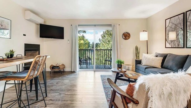 A living room with a sofa, a dining table with chairs, a TV mounted on the wall, and a balcony with a railing.