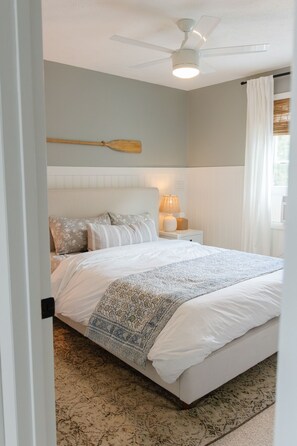 Quarto com cama, ventilador de teto, mesa de cabeceira com abajur e janela com cortinas.