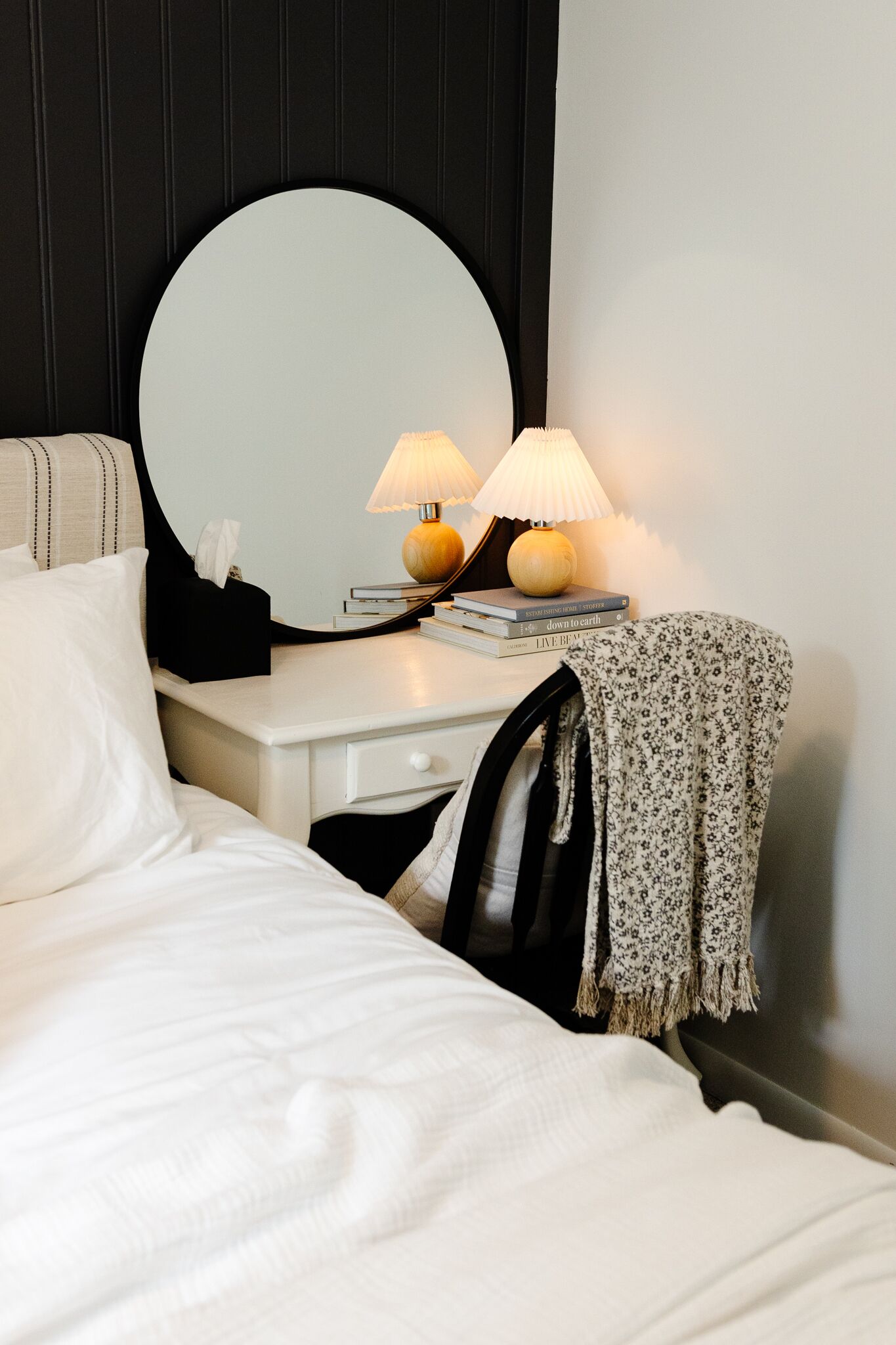 A neatly made bed with white linens, a bedside table with a lamp, a mirror, and a few books.