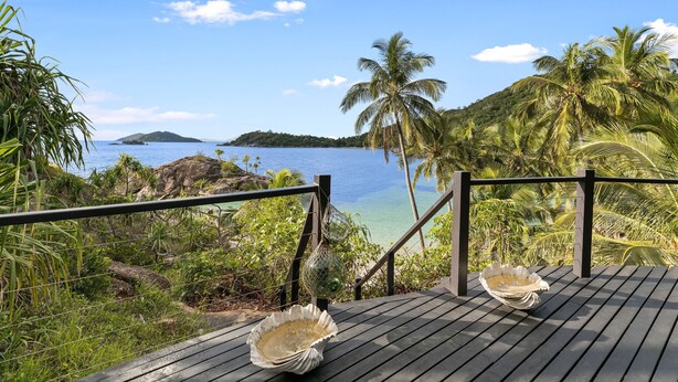 Une terrasse en bois ornée de deux coquillages, donnant sur une plage tropicale bordée de palmiers et baignée d’une eau d’un bleu cristallin.