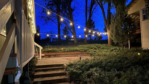 A residential backyard with a wooden staircase leading up to a deck, surrounded by lush greenery and string lights. - Near Yosemite & Lake & On Golf Course (Groveland)