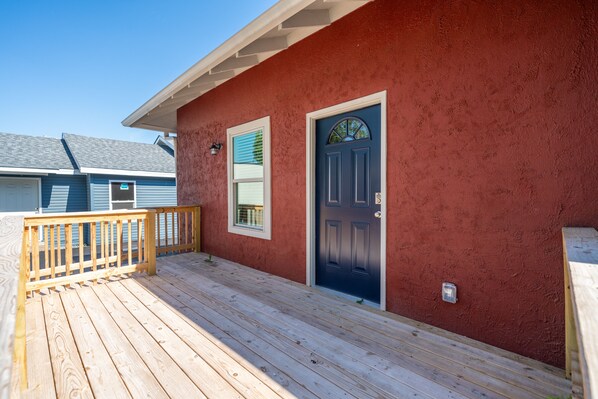 Una terraza de madera que conduce a una casa de color rojo, con una puerta azul y una ventana con persianas.