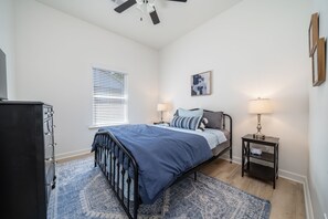 A bedroom with a bed, bedside tables, a dresser, a window with blinds, a ceiling fan, and a wall-mounted artwork.