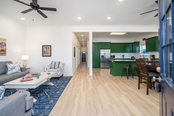 A modern living room with a sofa, coffee table, and a dining area with chairs and a table. A kitchen with green cabinets and a refrigerator is visible in the background.