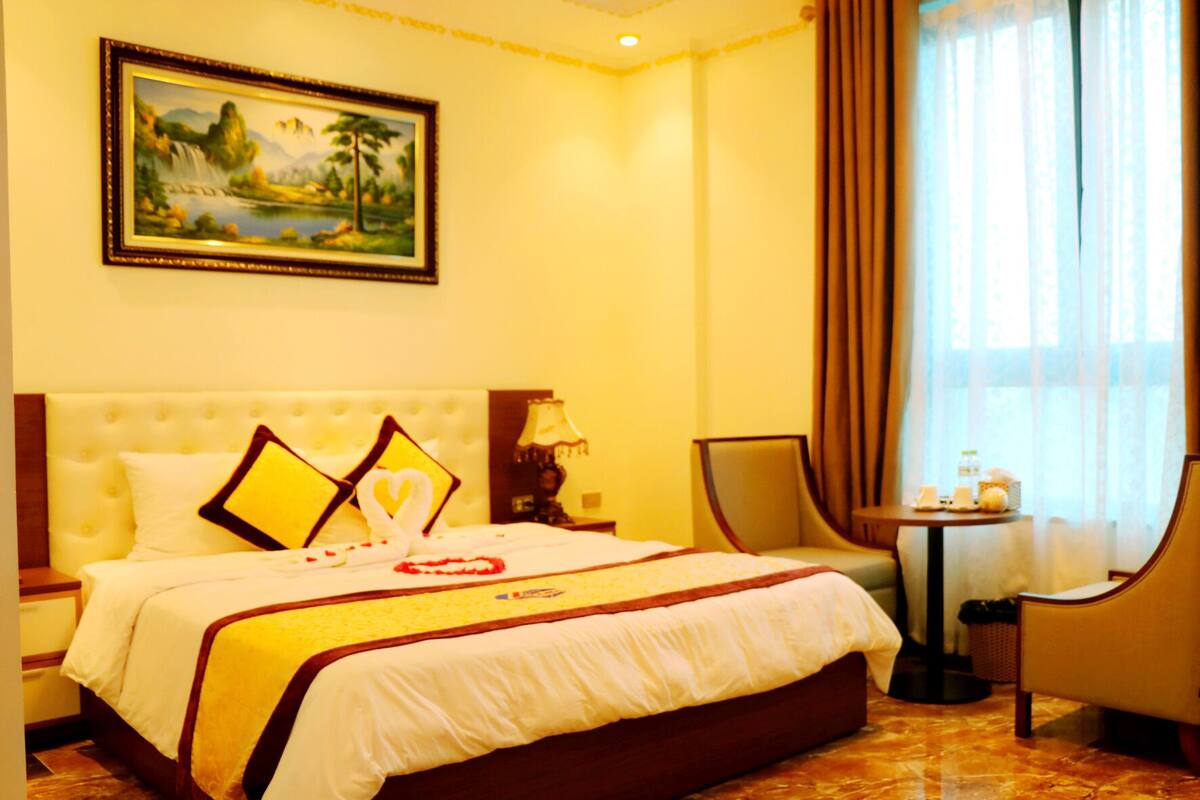 A hotel room with a large bed, a painting on the wall, a small table with a lamp, and a chair.