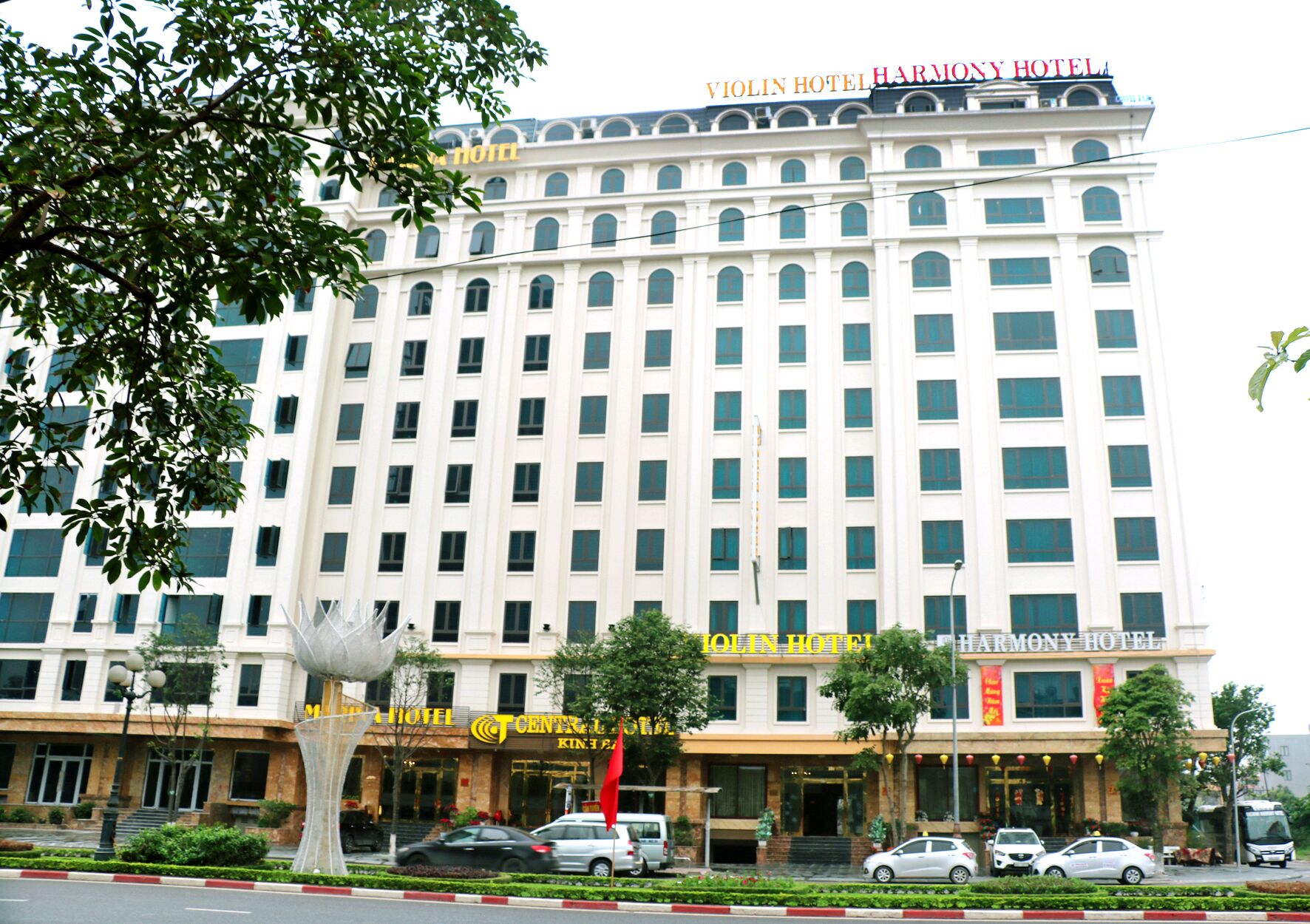 Un hotel de varios pisos con una entrada central, rodeado de vegetación y automóviles estacionados.