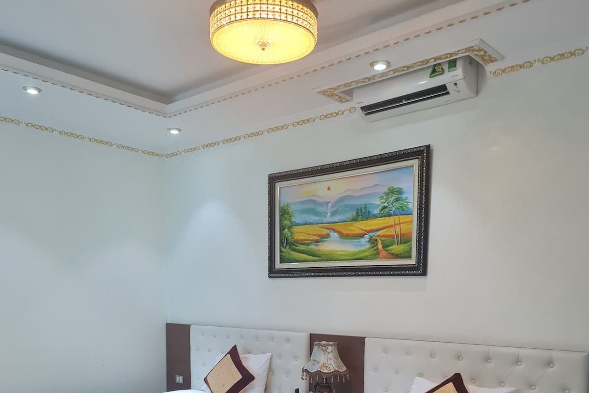 A hotel room with two beds, a ceiling light, an air conditioning unit, and a painting on the wall.