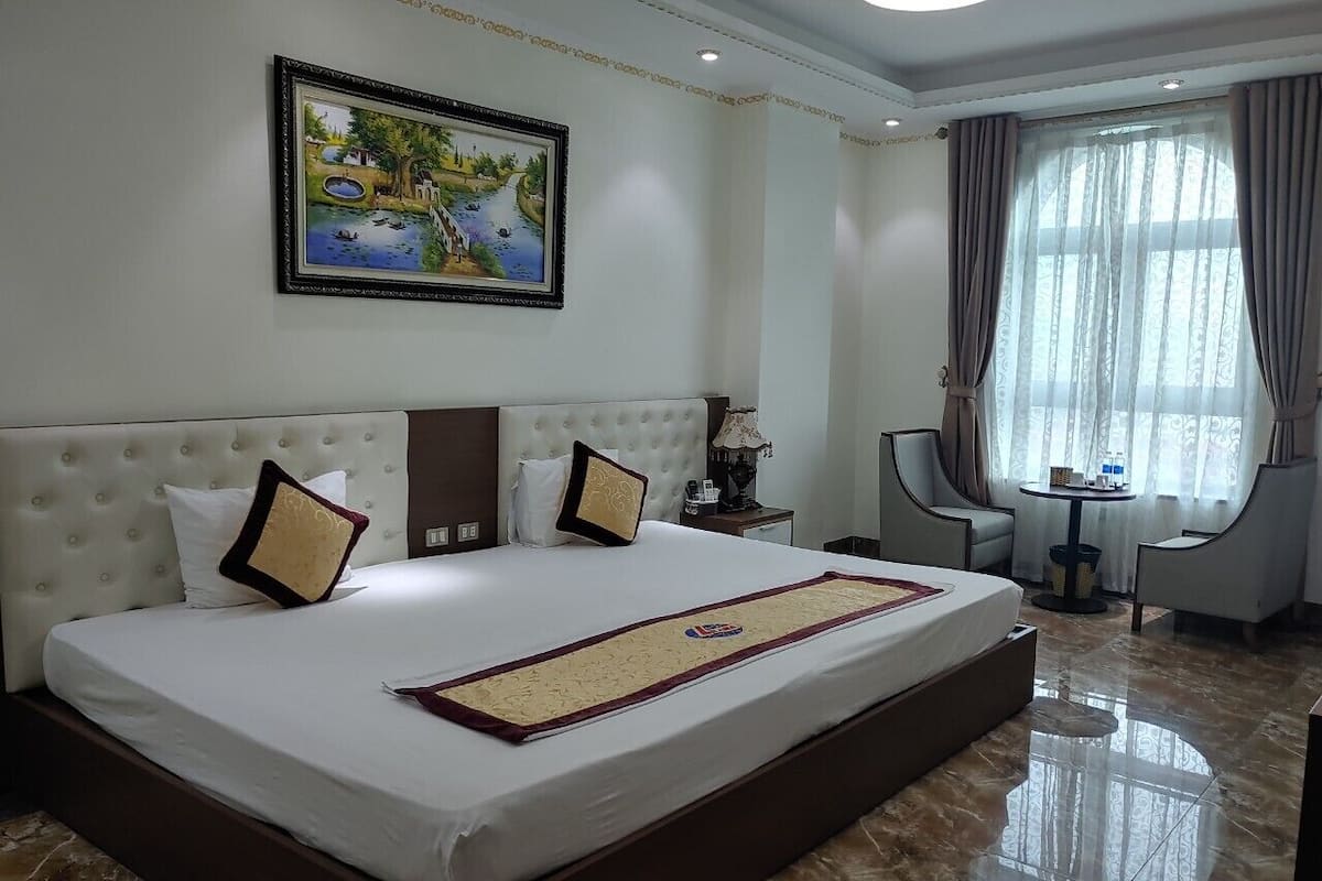 A hotel room with a large bed, a painting on the wall, a small table, and a chair.