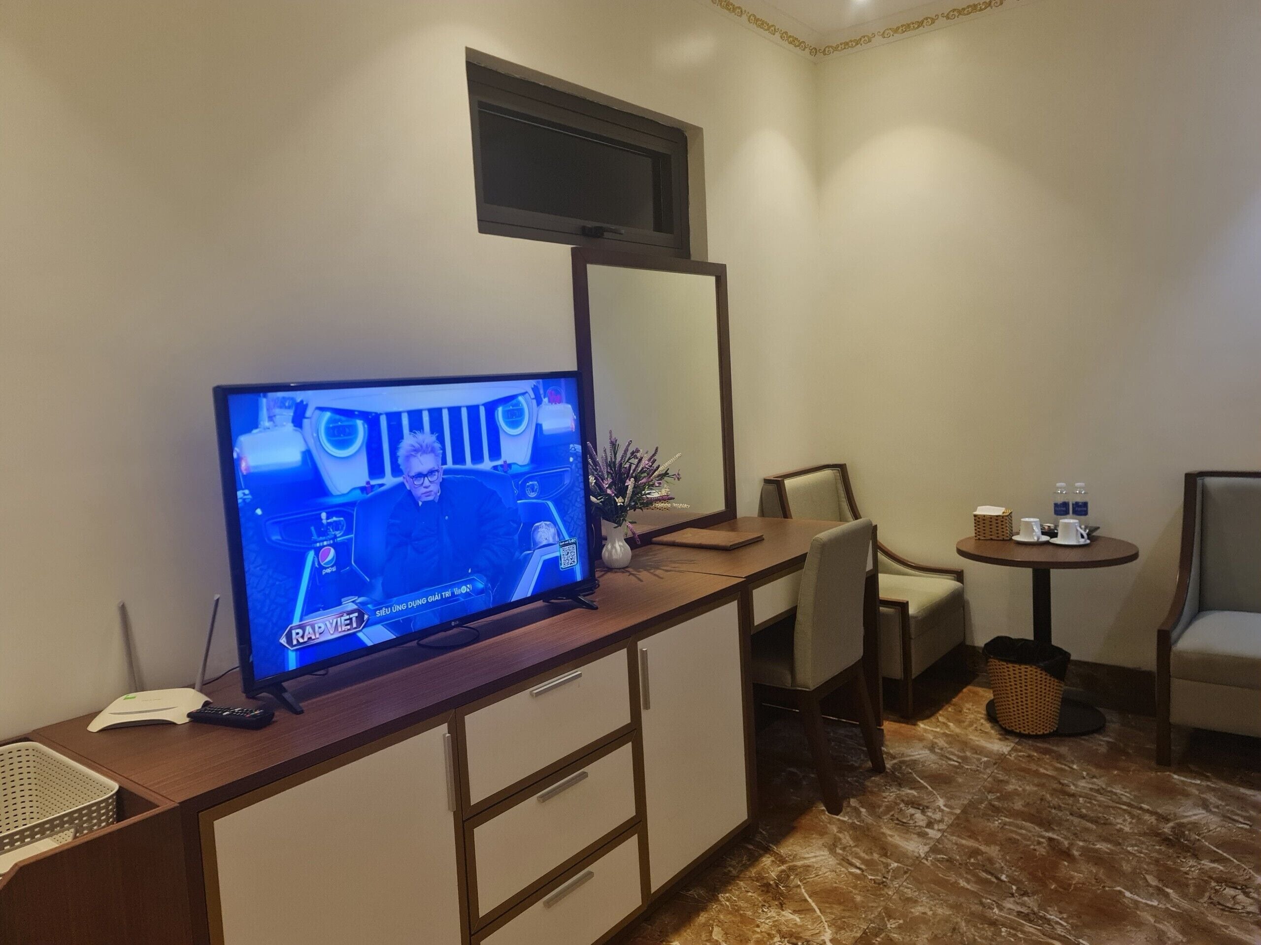 Habitación de hotel con televisor sobre un mueble de madera, una mesita redonda con un florero y una taza, y una silla.