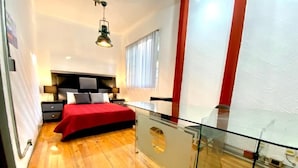 Un dormitorio moderno con suelo de madera, balcón de cristal, colcha roja y una planta grande en maceta.