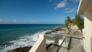 A balcony with a glass railing overlooking the ocean, a rocky shoreline, and a clear blue sky. - Stunning views of Seven Mile Beach, luxurious rooftop and oceanfront pool. (West Bay)