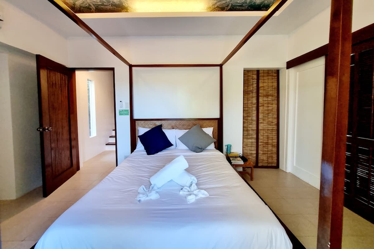 Une chambre d’hôtel avec un grand lit, une tête de lit en bois, une table de chevet avec une lampe et une fenêtre avec des stores.