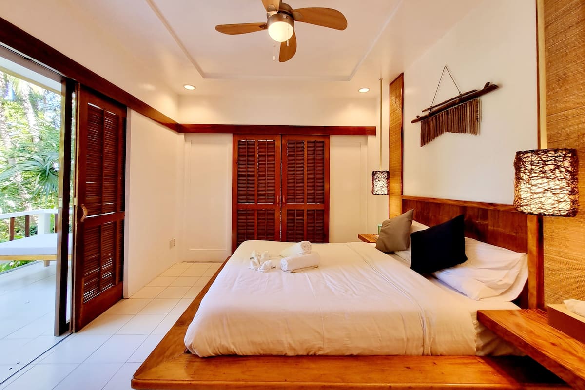 Une chambre à coucher avec un lit, une tête de lit en bois, un ventilateur de plafond et une vue sur l’extérieur.