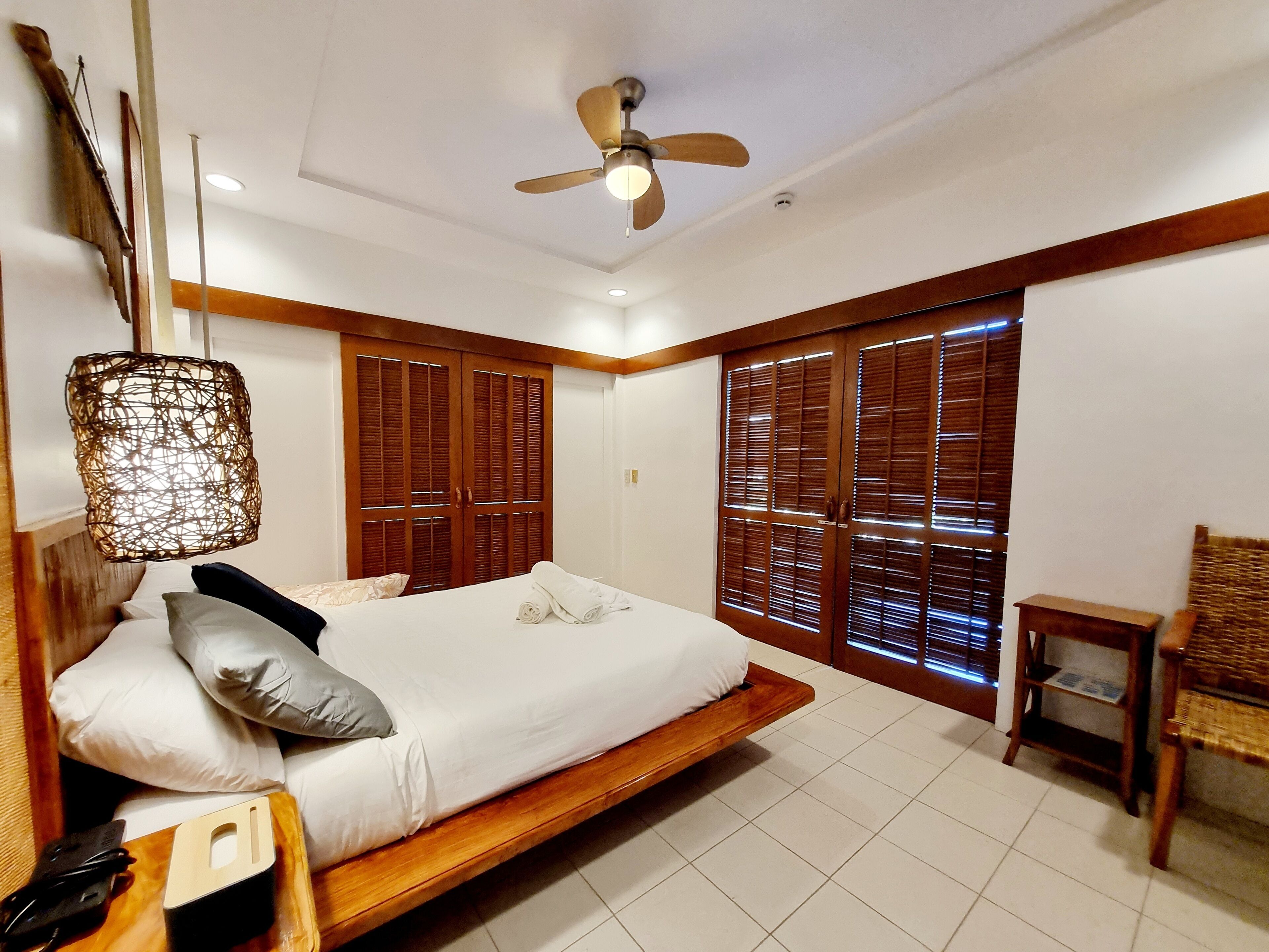 A bedroom with a bed, wooden wardrobe, ceiling fan, and a small table.