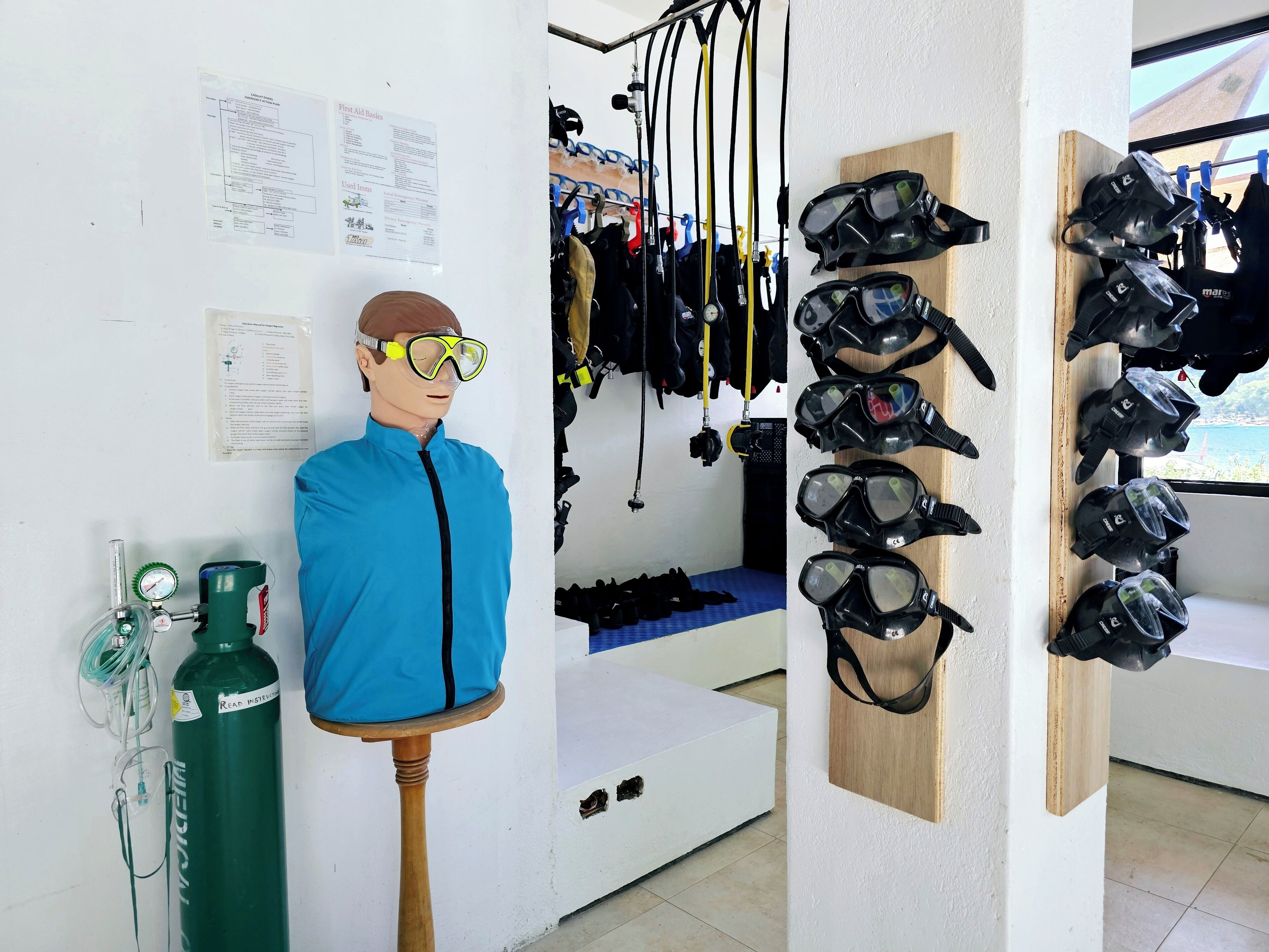 Una habitació amb un maniquí portant una jaqueta blava i ulleres grogues, un dipòsit d'oxigen verd, i diverses màscares i equips de submersió als prestatges.