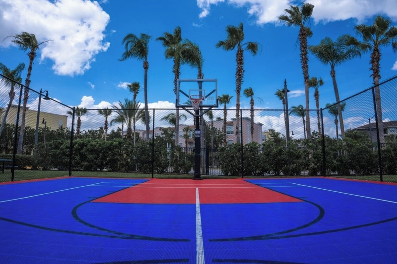 Een buitenbasketbalveld met een blauw en rood oppervlak, omringd door een hek en palmbomen.