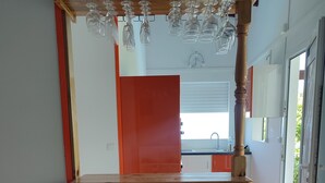 Eine Holzbar mit hängenden Weingläsern, einer roten Tür und Blick auf eine Küche sowie einen Außenbereich.
