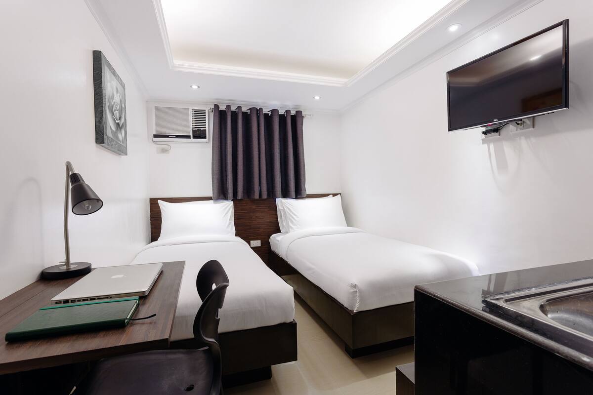 Una habitació d'hotel amb dos llits individuals, un escriptori amb un ordinador portàtil, una cadira, un televisor fixat a la paret i una finestra amb cortines.