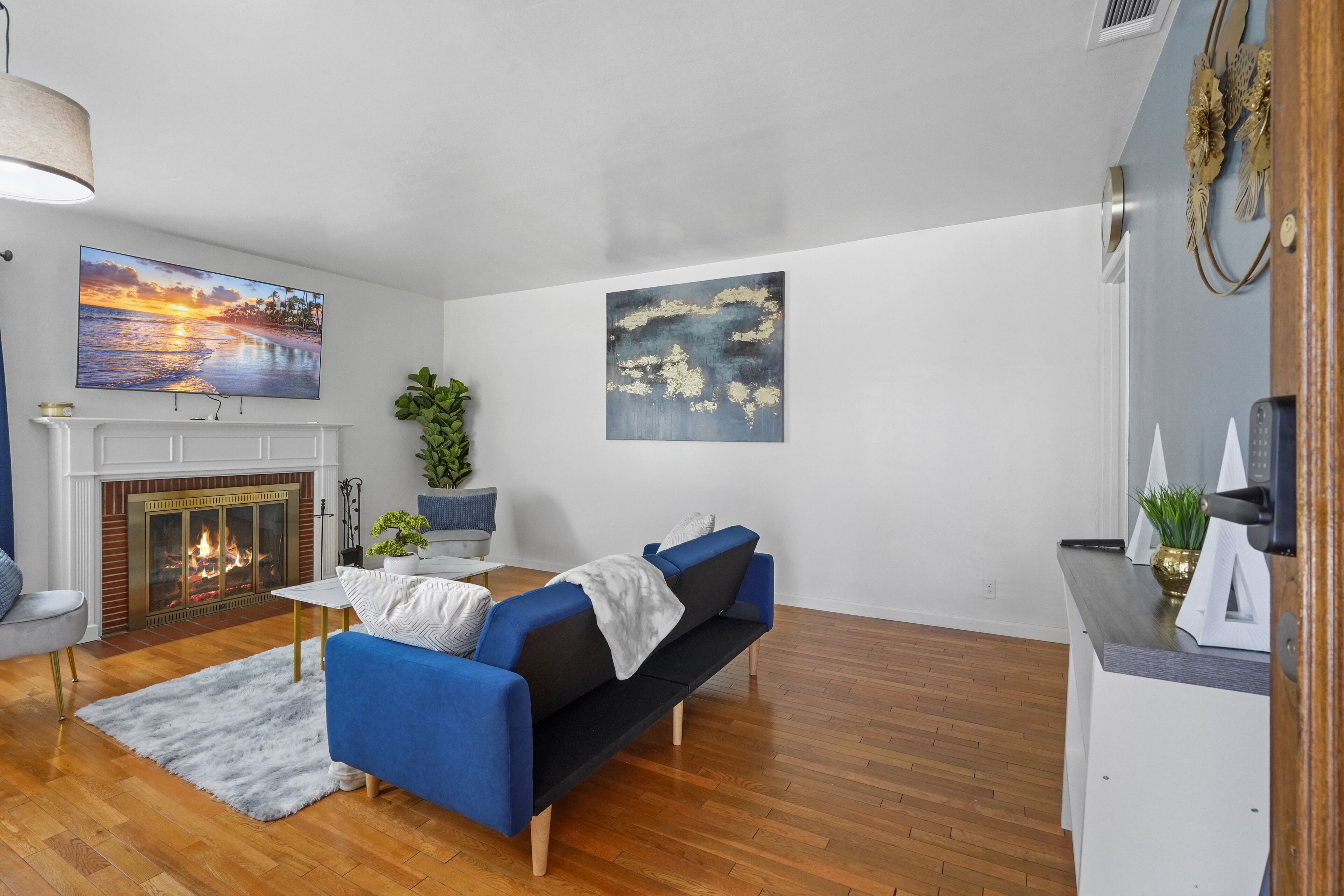 Een woonkamer met een open haard, een blauwe bank, een schilderij van een zonsondergang en een houten vloer.