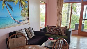 A room with a wooden dining table, a sofa with patterned cushions, a large beach-themed painting, and a view of the outdoors through large windows.